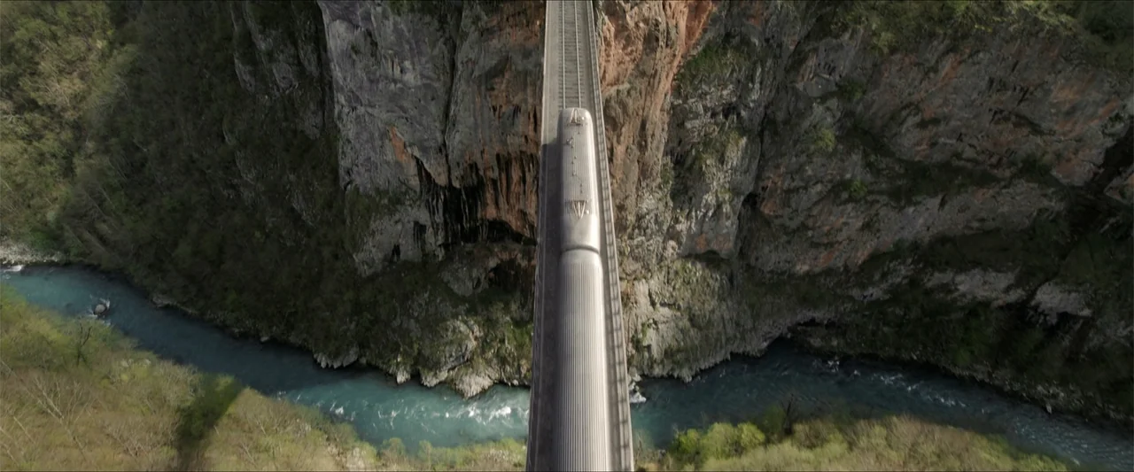 Heads of State (2025) movie still: establishing — A silver train travels across a narrow concrete bridge spanning a deep canyon. A blue river flows…