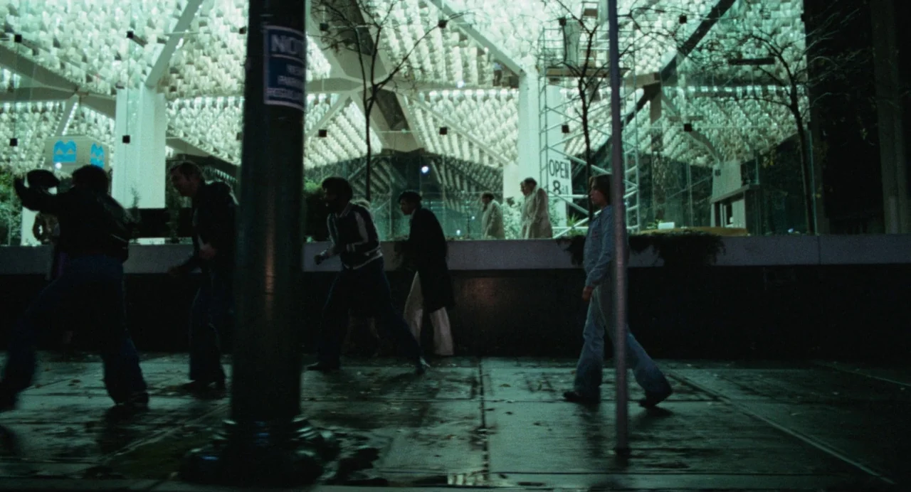 Out of the Blue (1980) movie still: wide — A dark city sidewalk at night runs in front of a modernist building facade. Multiple individuals walk…