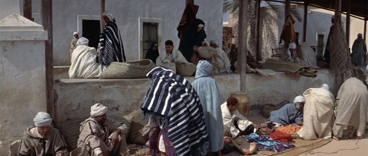 Legend of the Lost (1957) movie still: wide — An outdoor desert marketplace features low white walls and several people in traditional garments.…