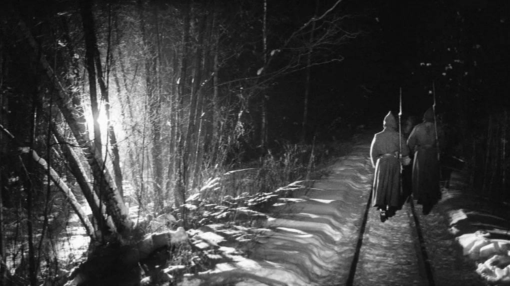 The Role (2013) movie still: wide — A snowy forest path at night runs alongside parallel railway tracks. Two soldiers wearing long overcoats and…