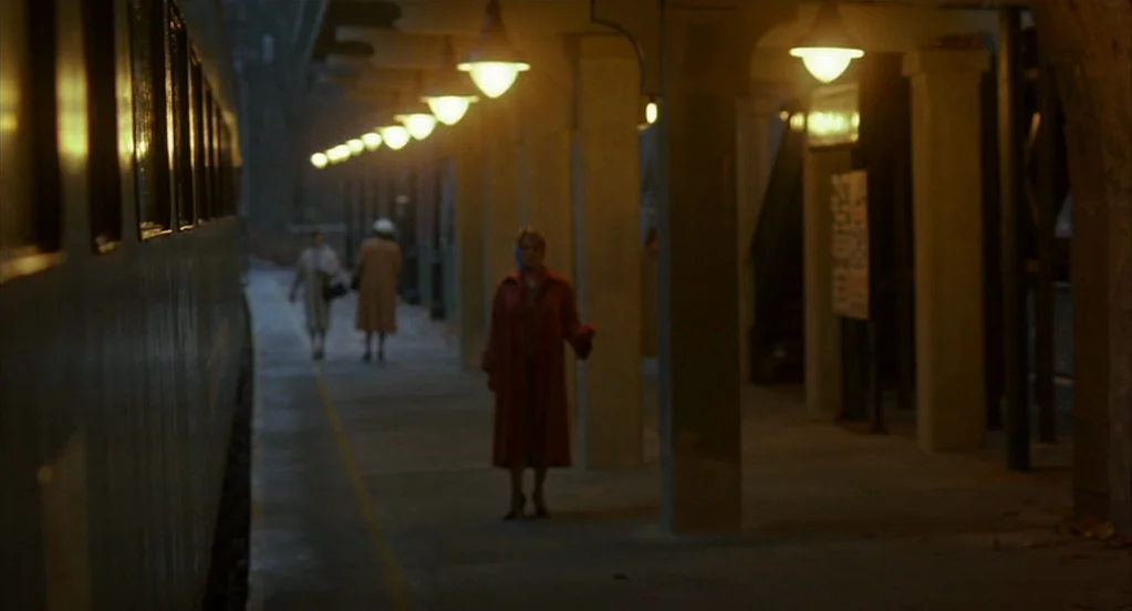 Far From Heaven (2002) movie still: wide — An dimly lit outdoor commuter train platform features concrete pillars and rows of hanging lamps. A woman…