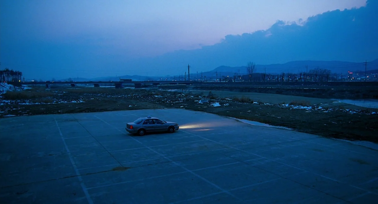 I Saw The Devil (2010) movie still: wide — A desolate concrete lot by a river is bathed in the cool blue twilight. A lone sedan rests in the center…