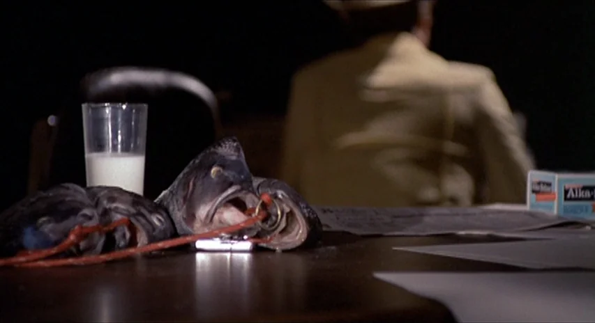 Blood Simple (1984) movie still: medium — A dark, dimly lit room focuses on a wooden table cluttered with peculiar objects. A person sits out of…