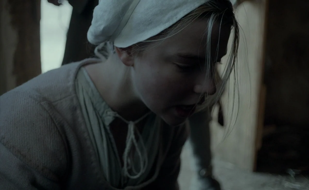 The Witch (2015) movie still: closeup — A dimly lit barn interior features wooden beams and rustic, primitive construction details. A young woman in…