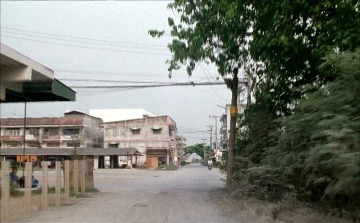 Blissfully Yours (2002) movie still: establishing — An unpaved dirt road stretches toward multi-story buildings under an overcast sky. Lush green…