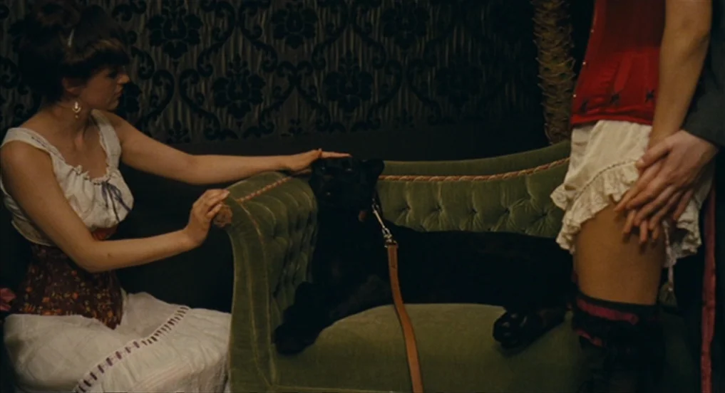 House of Tolerance (2011) movie still: medium — A dark parlor features damask wallpaper behind a plush green tufted velvet sofa. A woman in a corset…