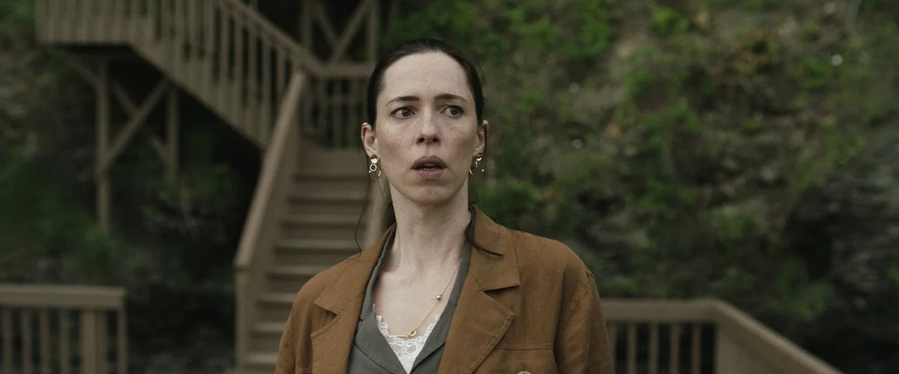 The Night House (2020) movie still: medium — A wooden staircase structure stands against a rocky hillside covered in sparse green vegetation. A…