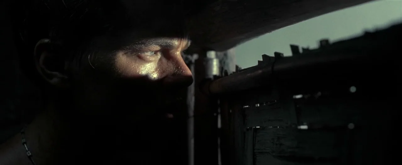The Beach (2000) movie still: closeup — A dark, cramped wooden enclosure with a narrow horizontal slat opening. A man peers through the gap with a…
