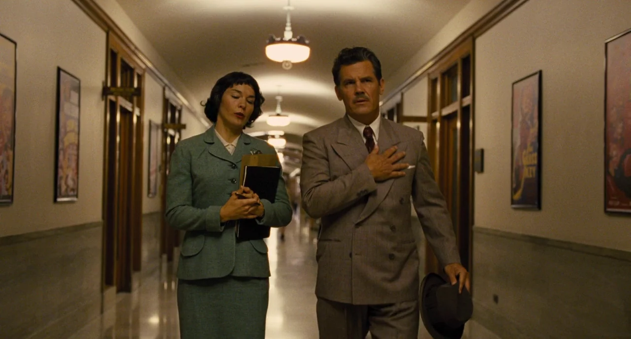 Hail, Caesar! (2016) movie still: fullbody — A long, symmetrical hallway in a 1950s office building with framed posters on walls. A man and a woman…