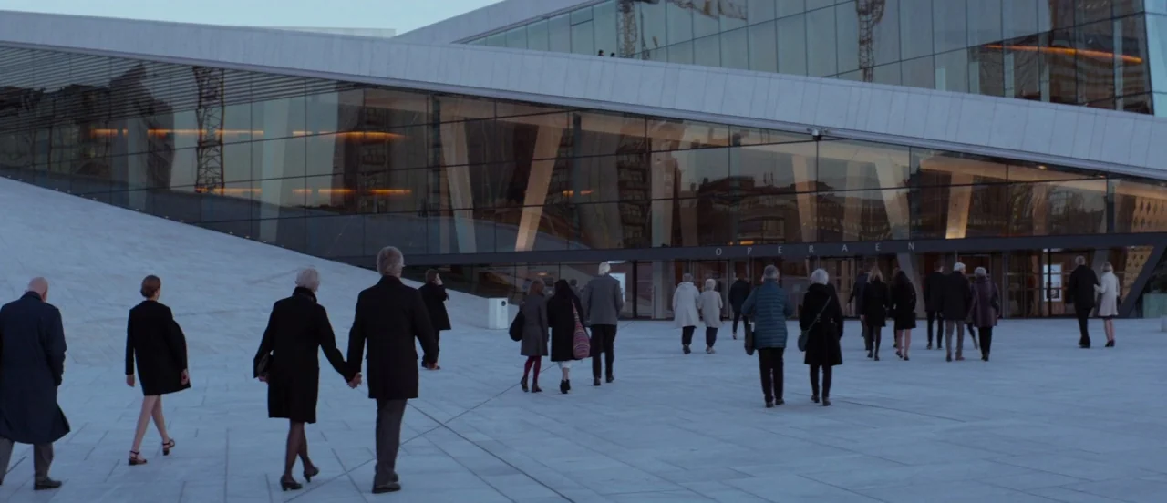 Thelma (2017) movie still: wide — A wide stone plaza leads toward the modern glass architecture of the Oslo Opera House. Pedestrians walk away from…