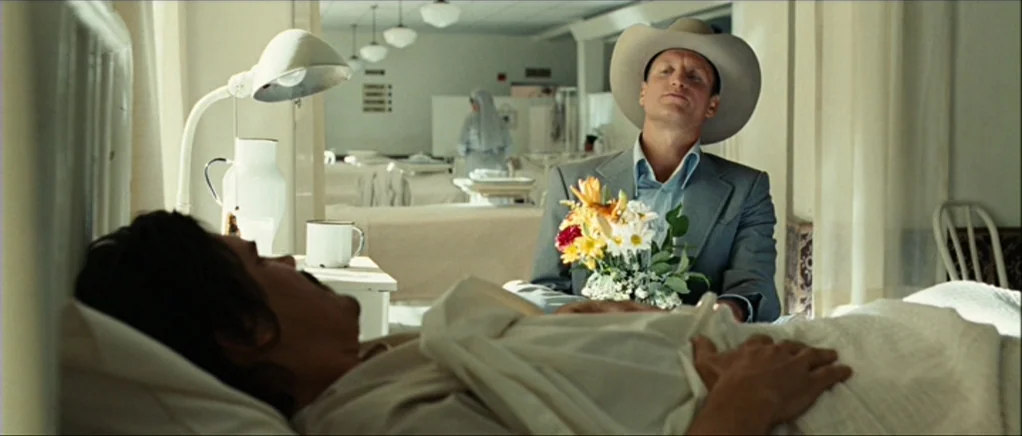 No Country For Old Men movie still: medium — A hospital ward with rows of white beds under uniform ceiling lights. A man in a cowboy hat sits next…