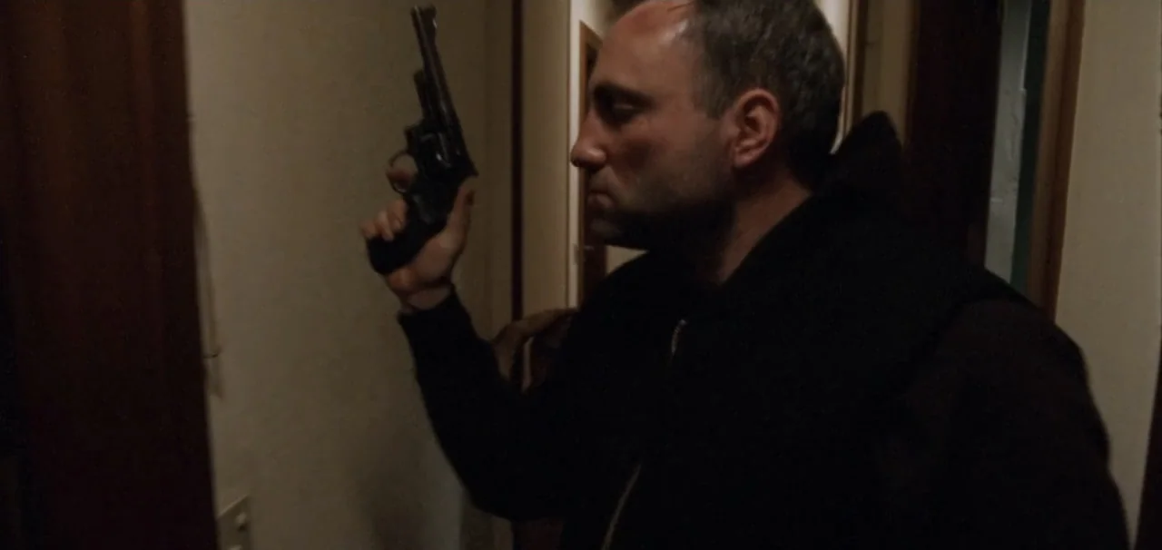 Pusher (1996) movie still: medium — A residential building hallway features beige walls and dark wooden trim under dim light. A man wearing a black…