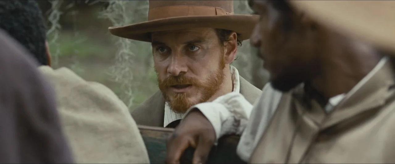 12 Years a Slave (2013) movie still: medium — An outdoor area with hanging foliage and a weathered wooden structure. A man with a red beard engages…