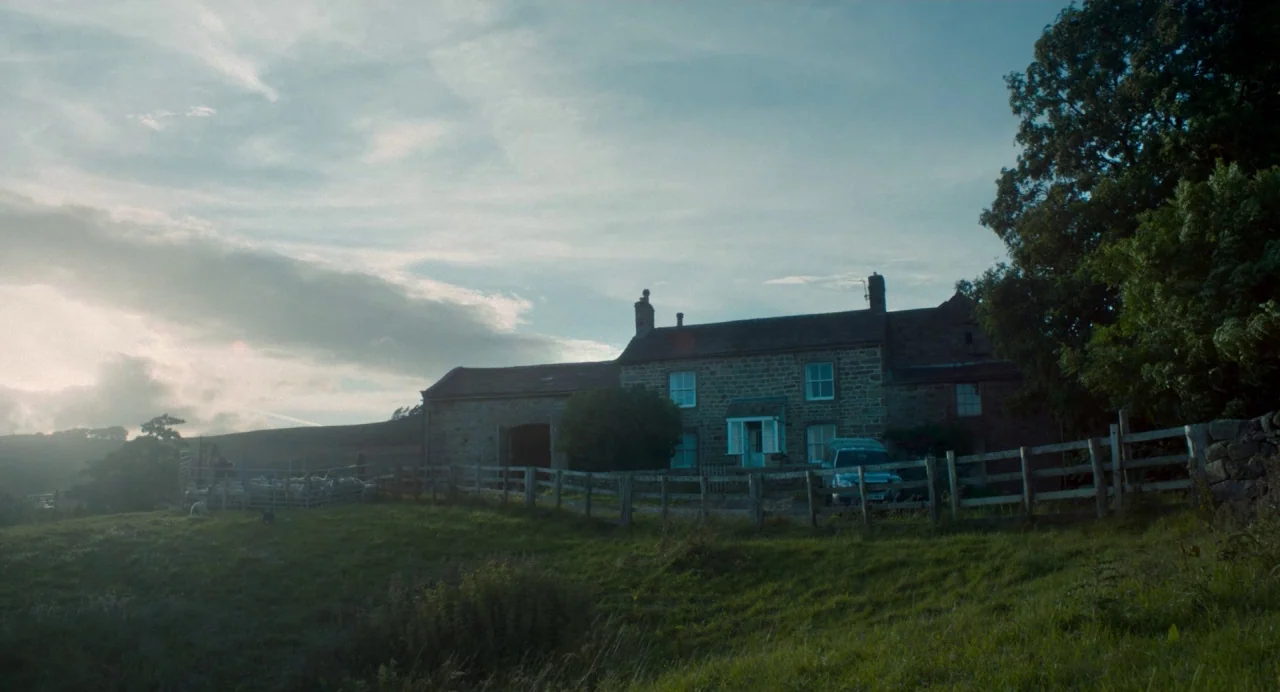 Dark River (2017) movie still: establishing — A stone farmhouse with a slate roof stands on a grassy slope under clouds. A wooden fence runs along…