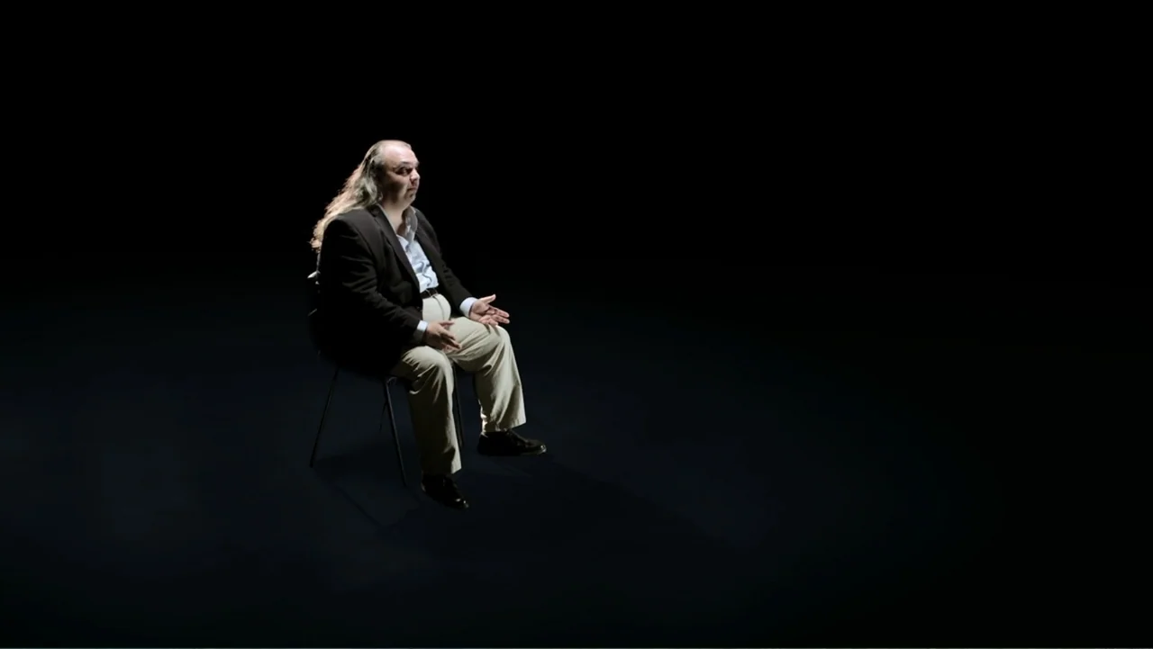Code of Trust (2019) movie still: medium — A person sits on a simple chair in a completely dark studio environment. The man rests on the chair while…