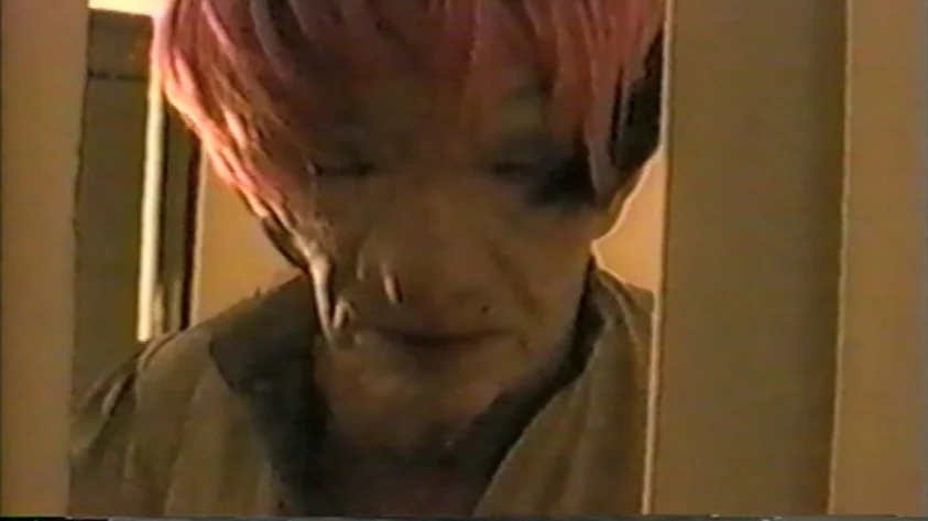 Trash Humpers (2009) movie still: closeup — An older man with a pink wig looks through a narrow vertical door opening. The frame exhibits strong VHS…