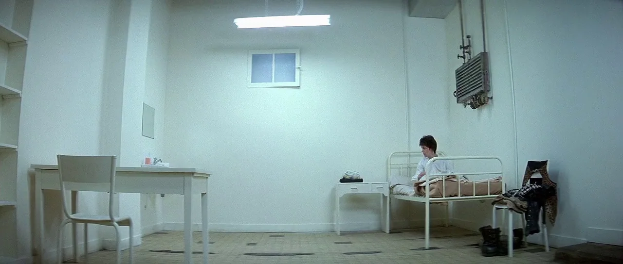 Nikita (1990) movie still: wide — A stark white, minimalist room resembles a clinical or institutional setting. A young man sits alone on a white…