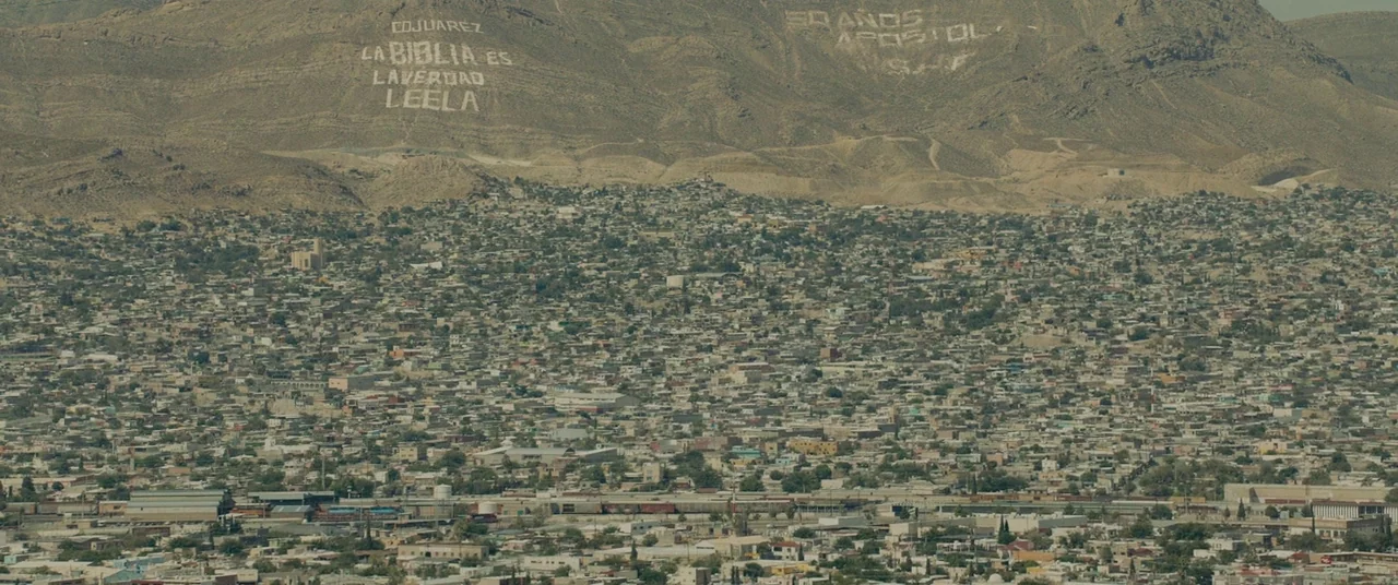 Sicario (2015) movie still: establishing — A dense urban sprawl stretches across a valley floor toward arid desert mountains. White text is painted…