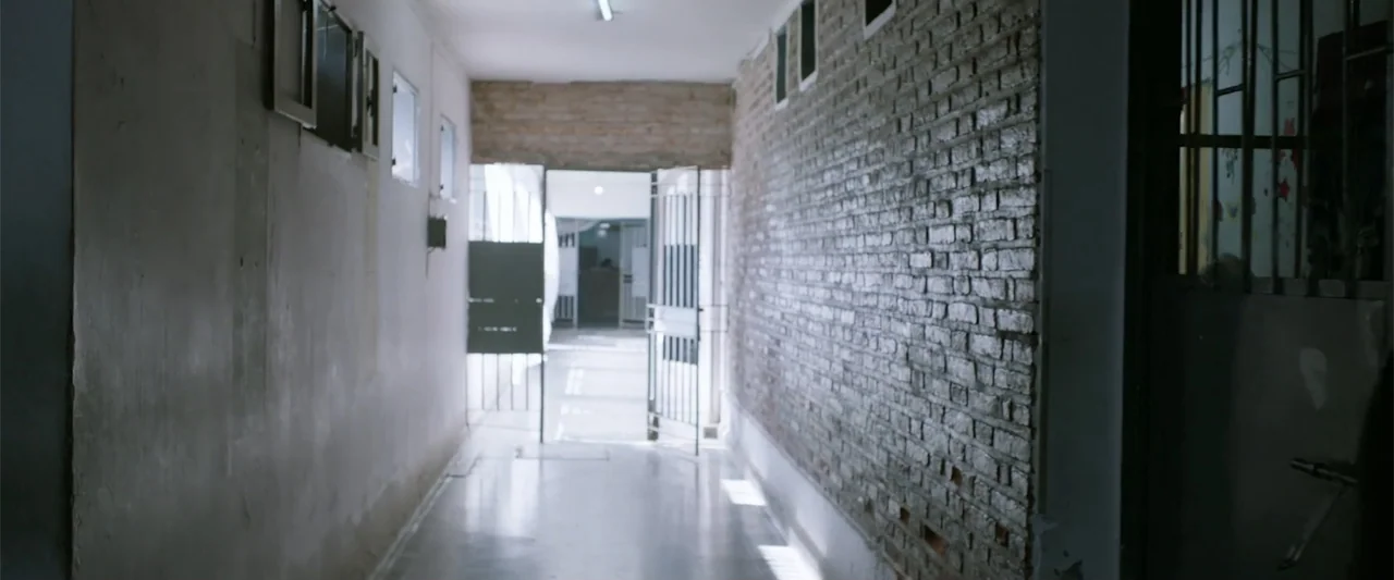 I Trust in You (2016) movie still: establishing — A narrow institutional hallway features a smooth concrete floor and a bare brick wall. An open…
