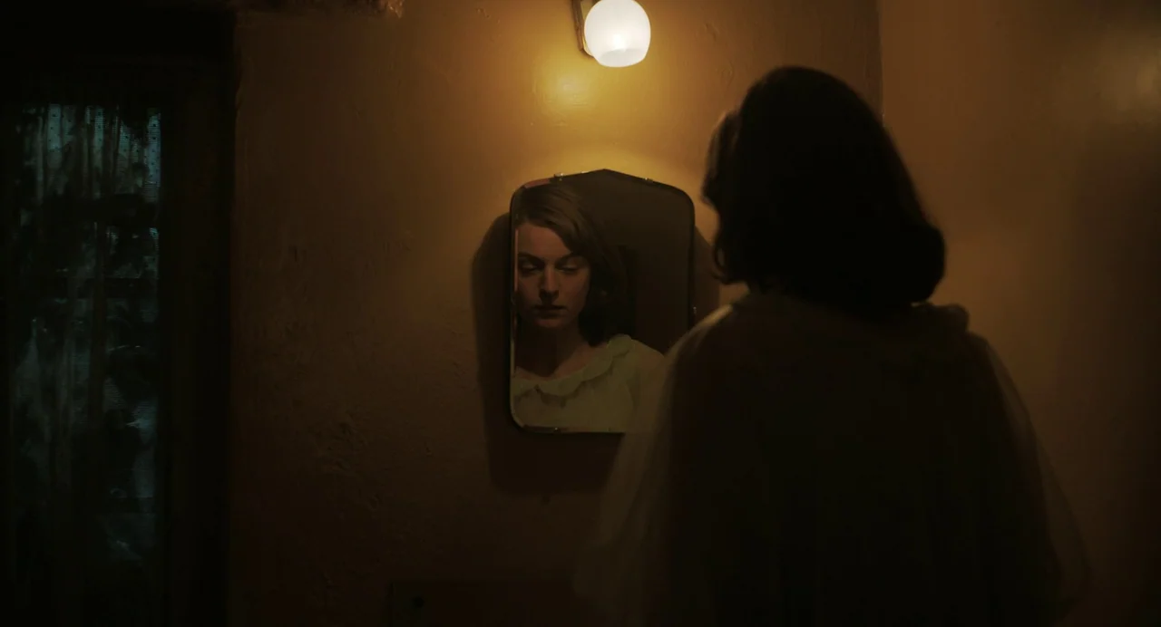 My Policeman (2022) movie still: medium — A dimly lit room with textured plaster walls and a single mirror. A woman in a dressing gown stands before…