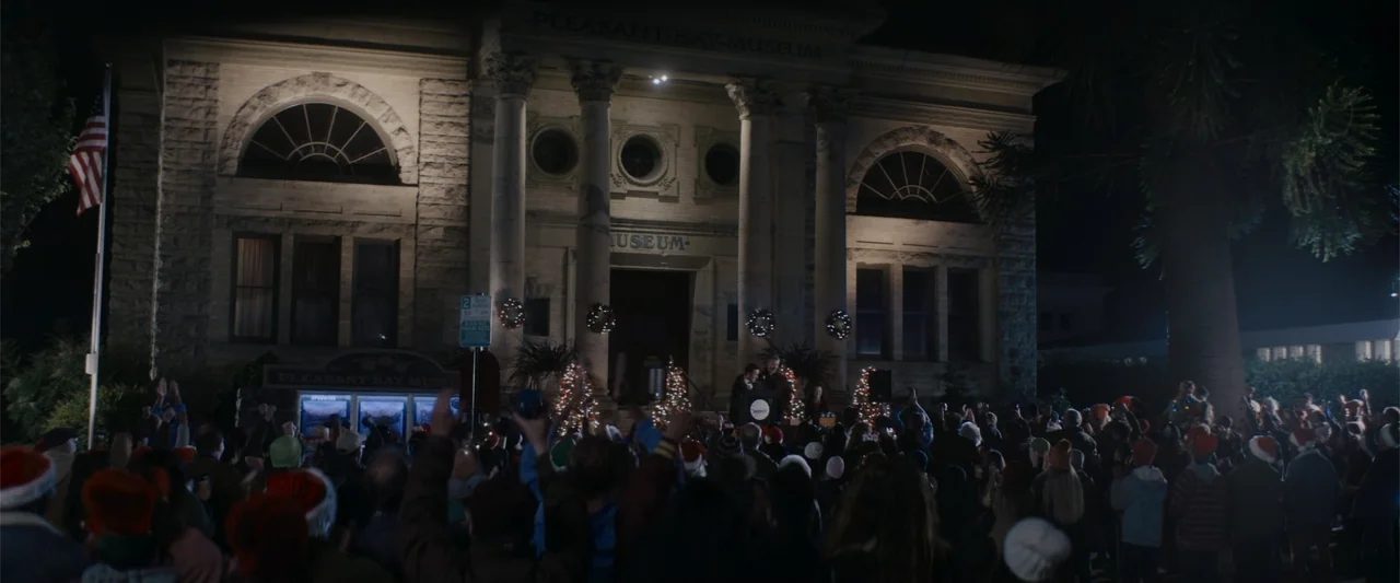 A Christmas Mystery (2022) movie still: wide — A stone museum building with classical columns serves as the backdrop for a crowd. Many people stand…