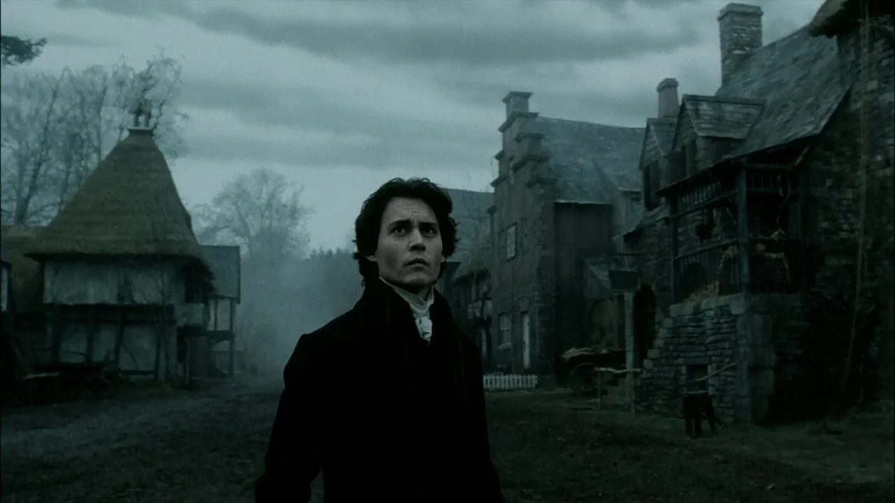 Sleepy Hollow (1999) movie still: medium — A misty, desolate village features stone cottages with thatched roofs under a cloudy sky. A man in a dark…