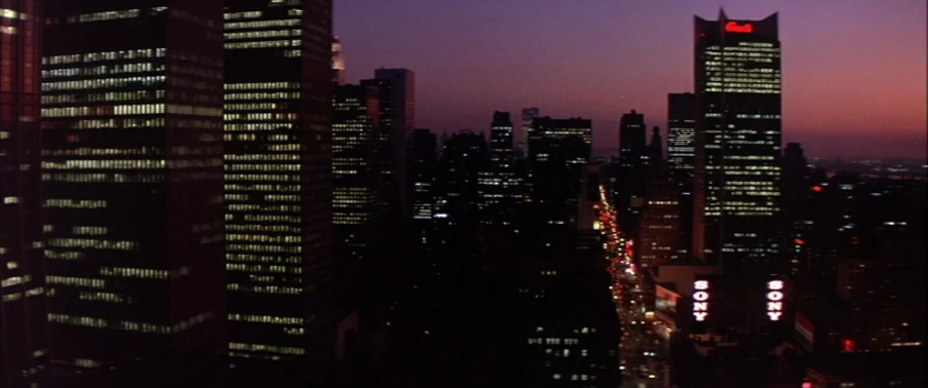 The Man Who Fell To Earth (1976) movie still: establishing — A dense urban skyline at dusk shows multiple illuminated office towers against a…