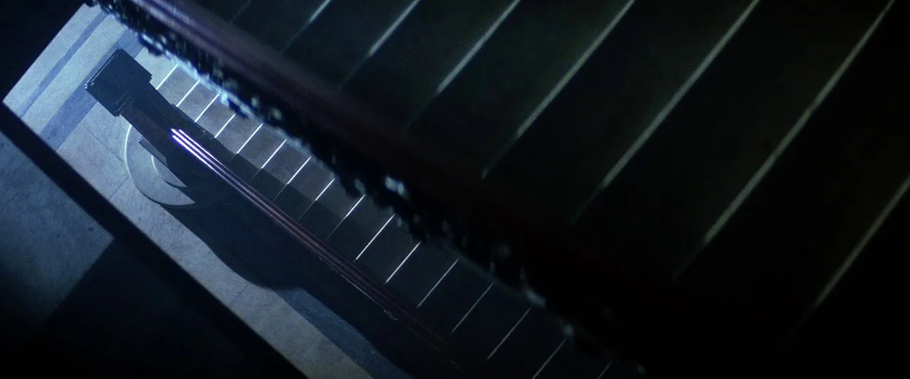 The Hunger (1983) movie still: wide — A high-angle view looking down a straight wooden staircase into darkness. Sharp, linear highlights cut through…