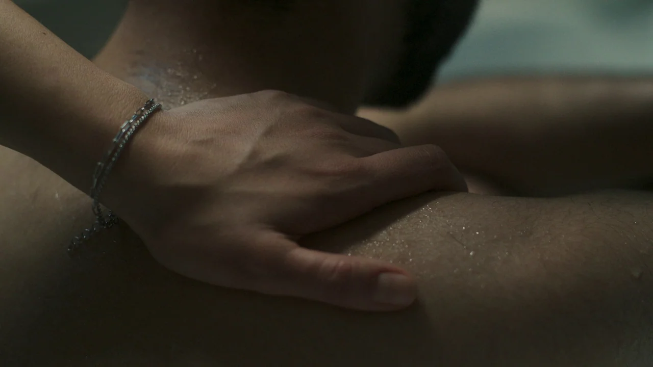 Helll Hole (2024) movie still: closeup — A dark, moisture-laden surface shows the skin of a shoulder and a hand. A person's hand with a metallic…
