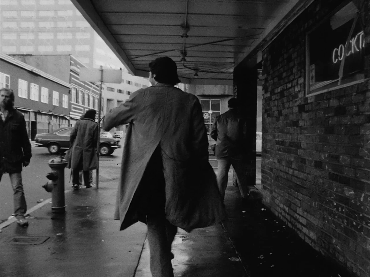 Mala Noche (1986) movie still: fullbody — A city sidewalk beside a brick building featuring a neon cocktails sign. A man in a long coat walks away…