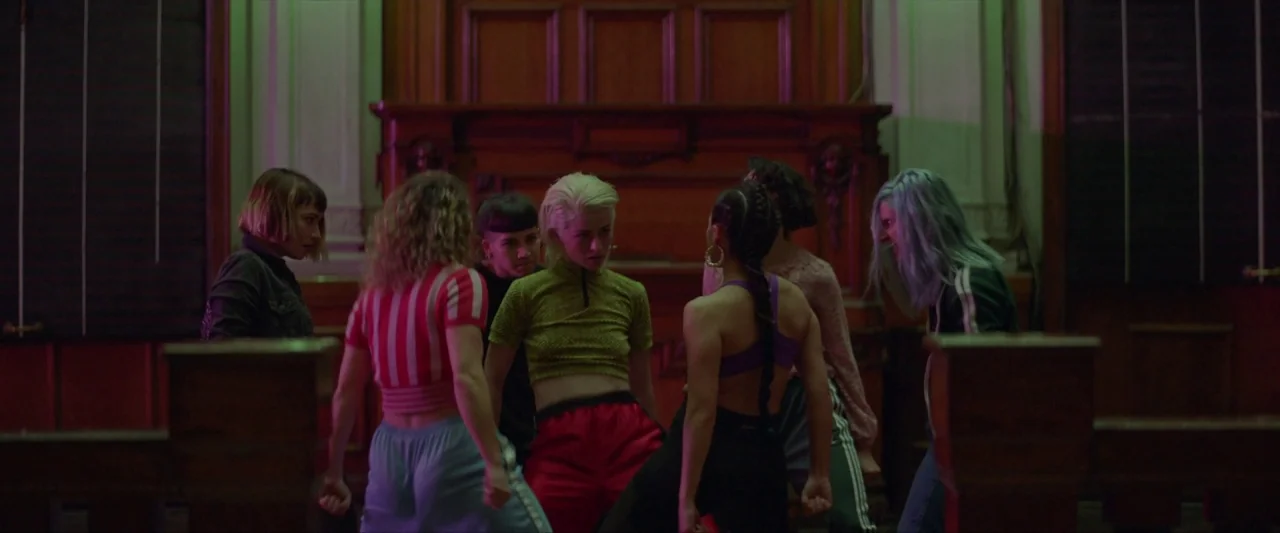 Ema (2019) movie still: medium — A dark, wood-paneled ballroom features ornate architectural details and a large empty fireplace. Six women in…