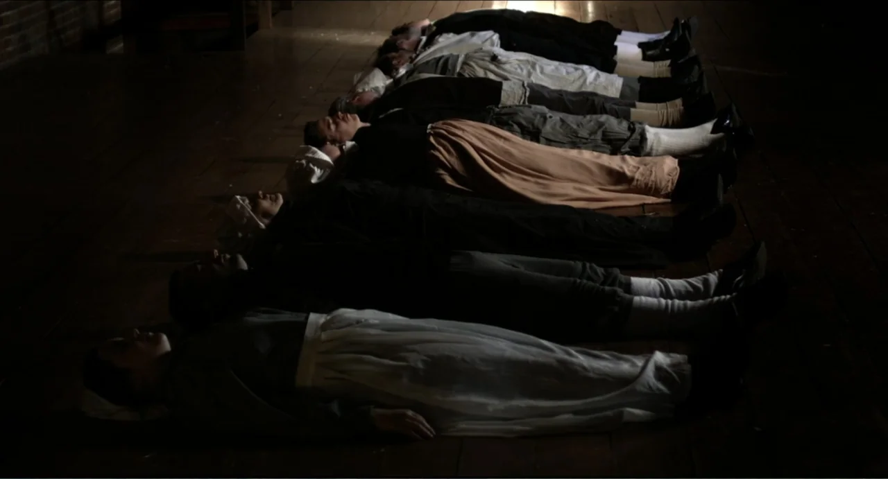 The Secret Village (2013) movie still: fullbody — A row of individuals in period clothing lie aligned on a dark wooden floor. They are resting or…