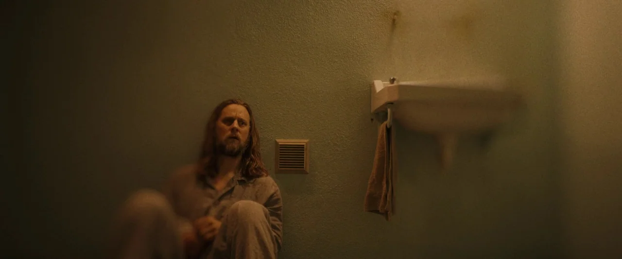 Oddity (2024) movie still: medium — A sterile, dimly lit cell features a plain green wall with a small vent. A man with long hair sits on the floor…
