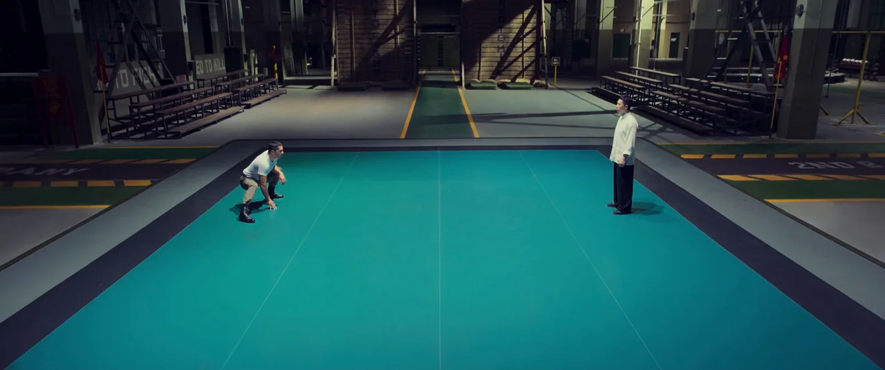 Ip Man 4: The Finale (2019) movie still: wide — A vast industrial training hangar features a large teal matted surface in the center. Two men stand…