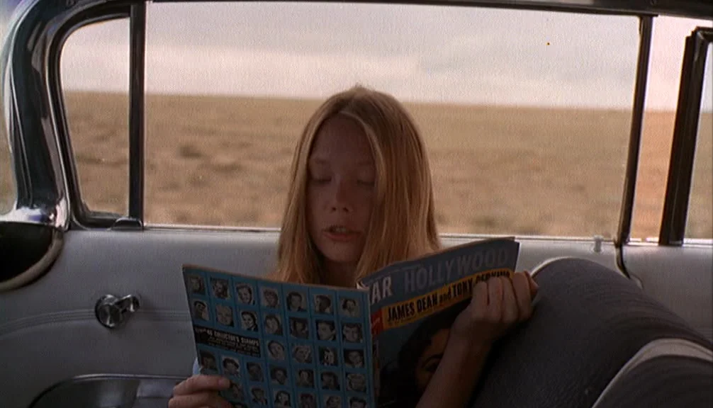 Badlands (1973) movie still: medium — A teenage girl sits in the back of a car in a desert landscape. She holds a magazine in front of her face…