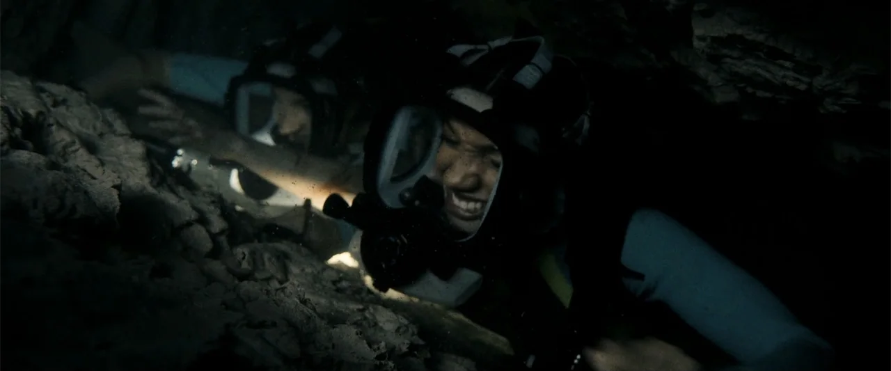 Anaconda (2024) movie still: medium — A dark, cramped underwater cavern features rocky textures and murky, light-deprived water. Two divers navigate…