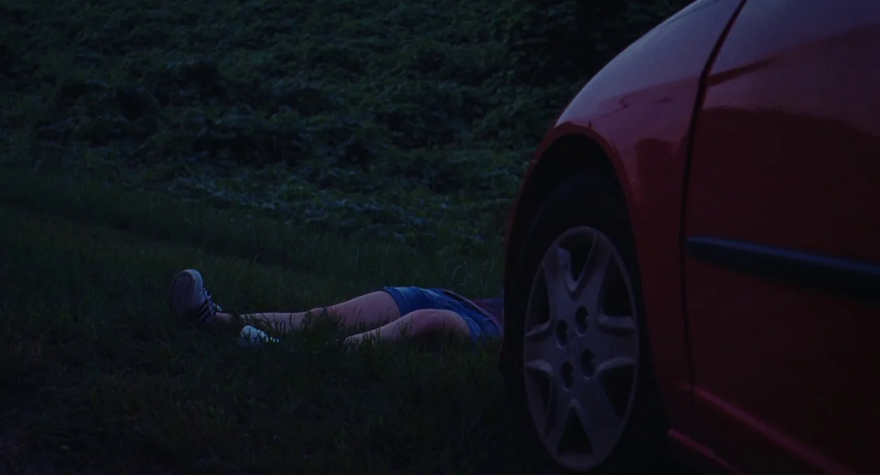The Giant (2019) movie still: wide — A dark grassy area sits beside the large red wheel of a parked car. A person lies horizontal on the ground…