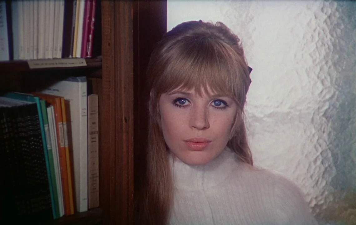 The Girl on a Motorcycle (1968) movie still: closeup — A woman stands beside a wooden bookshelf packed with various books and colorful spines. She…