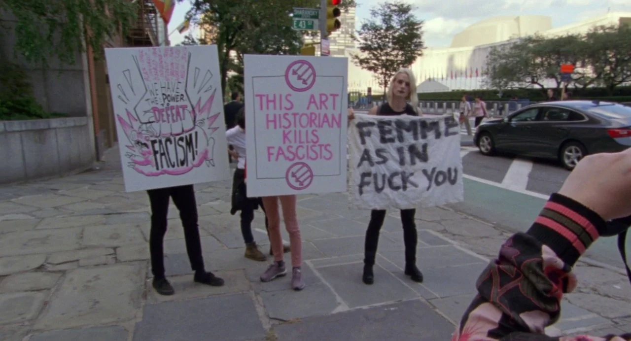 So Pretty (2019) movie still: fullbody — A group of people hold protest signs on a paved sidewalk near a street corner. The signs display…