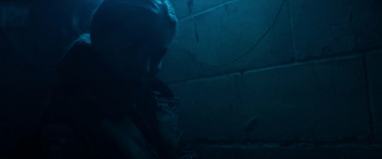 The Silencing (2020) movie still: medium — A dark, cramped room featuring a cinder block wall textured by mortar lines. A woman in a dark jacket…