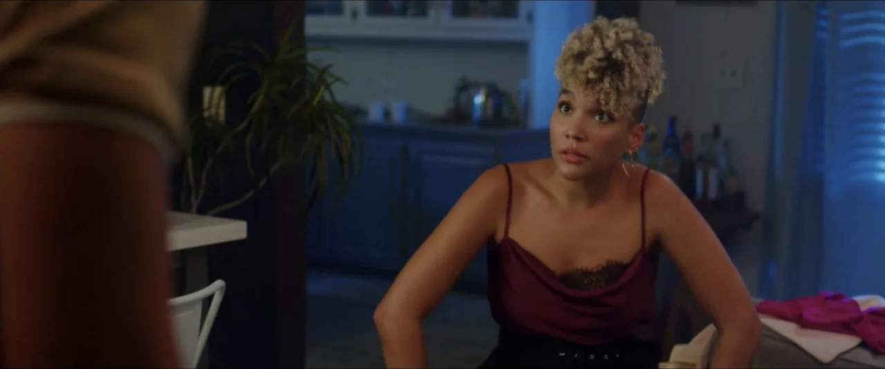 Gatlopp: Hell of a Game (2022) movie still: medium — A dimly lit room with a blue-toned color palette and modern furniture. A young woman with curly…