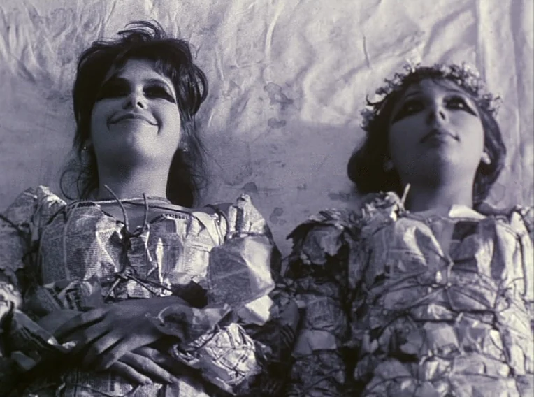 Daisies (1966) movie still: medium — Two women wearing dresses constructed from newspapers lie on the grass. They look directly upward while…