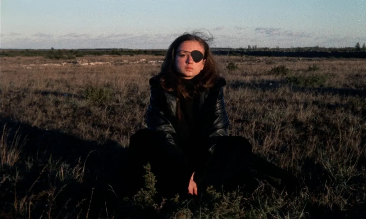 Thriller: A Cruel Picture (1973) movie still: medium — A flat, grassy field stretches toward a distant horizon under a clear sky. A woman wearing an…