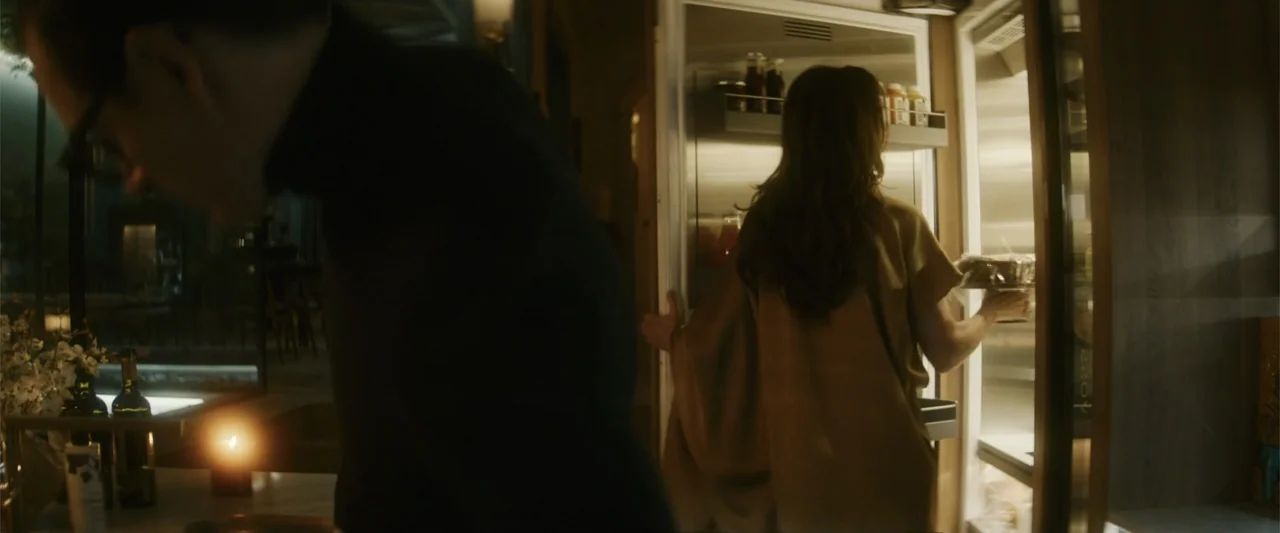 Black Bag (2025) movie still: medium — A dimly lit room features a glowing open refrigerator illuminating a person in tan. A man wearing a dark…
