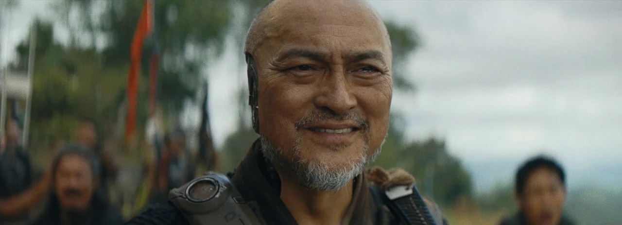 The Creator (2023) movie still: closeup — An older East Asian man with a robotic ear component stands in an outdoor area. He stares slightly…