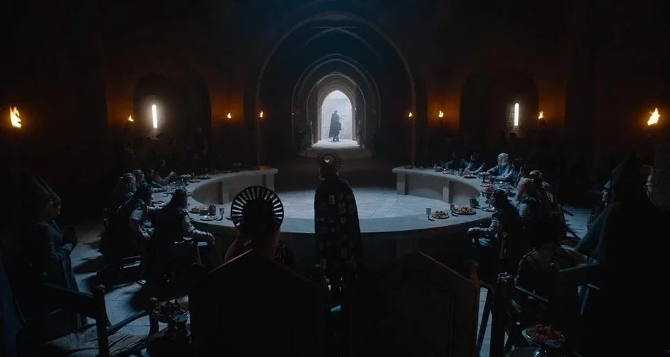The Green Knight (2021) movie still: wide — A circular stone hall features a large table surrounded by people in dark garments. A lone silhouetted…