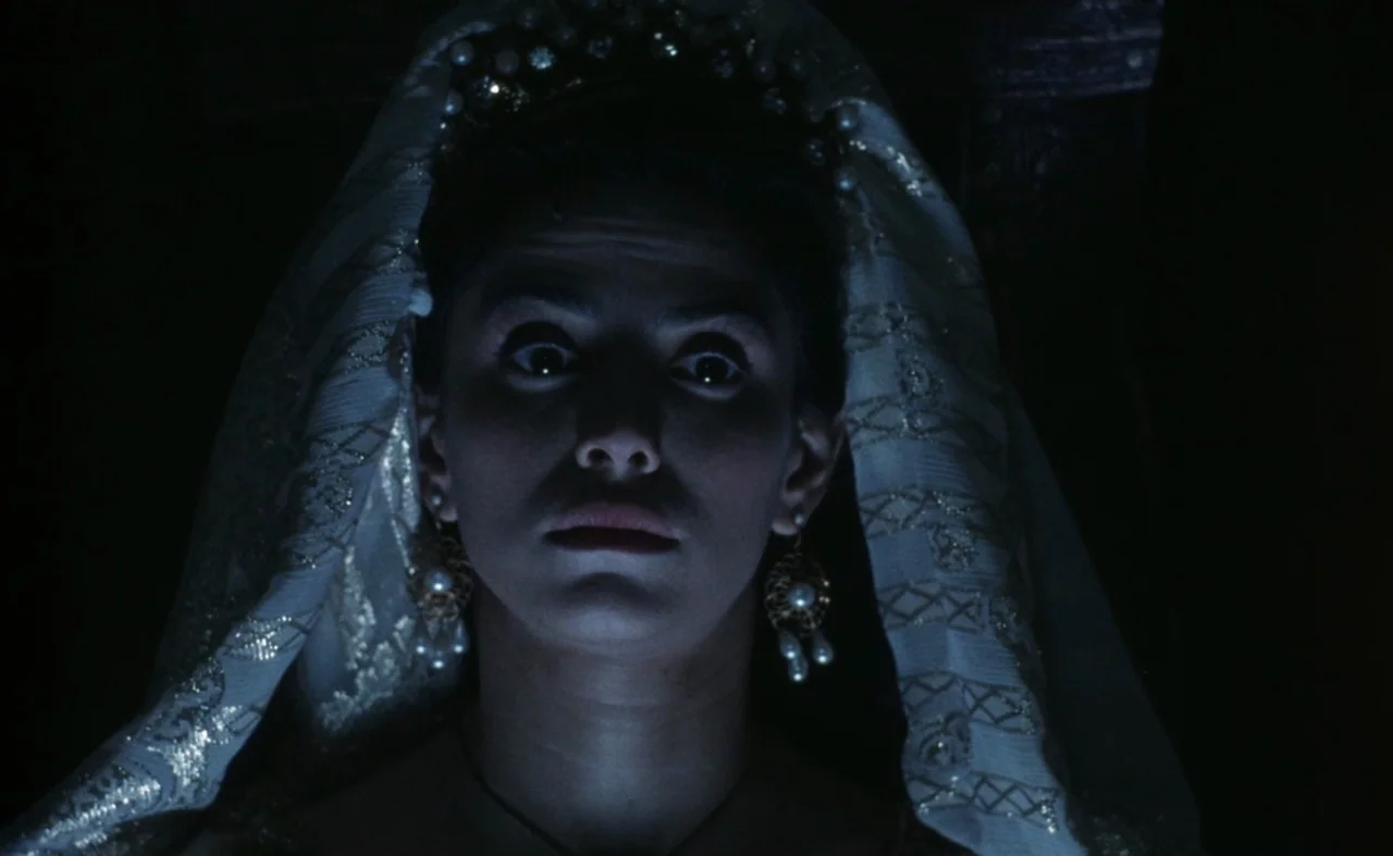The Pit and The Pendulum (1991) movie still: closeup — A woman wears an elaborate lace headpiece and ornate pearl jewelry in darkness. Her wide-eyed…
