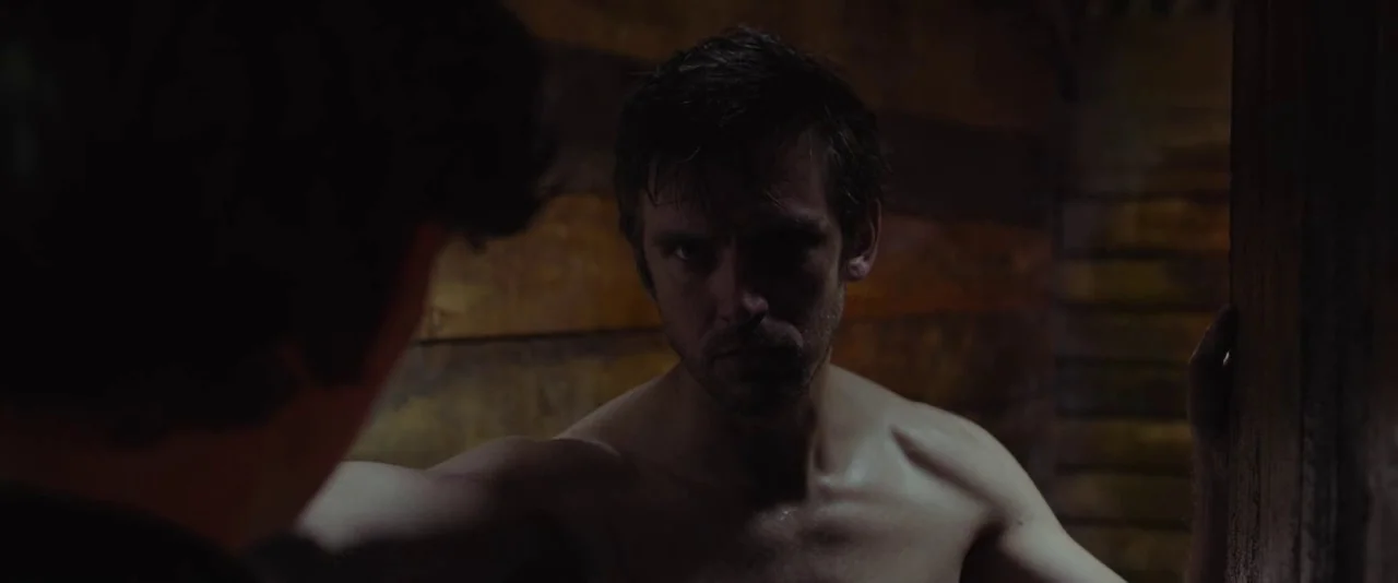 Apostle (2018) movie still: medium — A dimly lit room features walls constructed from rough-hewn wooden planks. A shirtless man stands facing…