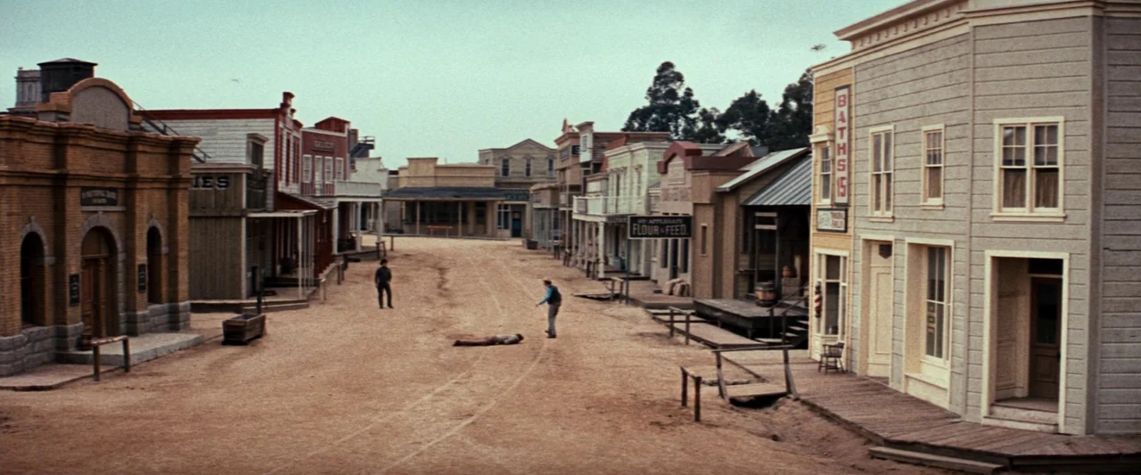 Westworld (1973) movie still: establishing — A dusty dirt street in a quiet frontier town lined with wooden storefronts. Three figures are spaced…