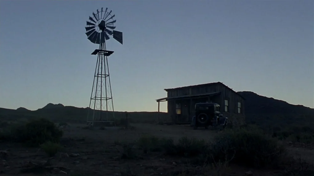 Ask The Dust (2006) movie still: wide — A dark, rustic wooden shack stands in a barren desert at dusk. A tall metal windmill and an old automobile…